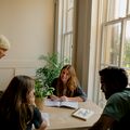 Four people collaborating at a table with notebooks and a plant, next to a window with natural light.