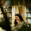 A person with curly hair is sitting in a library, wearing headphones and using a laptop. Bookshelves are in the background.