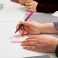 Lähikuva käsistä kirjoittamassa vaaleanpunaisella kynällä muistilapulle valkoisen pöydän ääressä, taustalla muita lappuja ja ihmisiä.