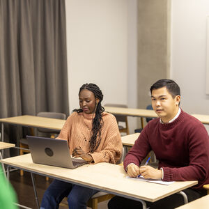 Two students sit in the front row, one looking at the teacher and the other at a computer.