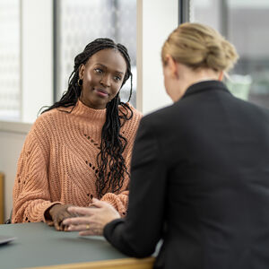 Two people having a conversation in an office setting, one wearing a peach sweater, the other in a black jacket. A laptop is on the table.