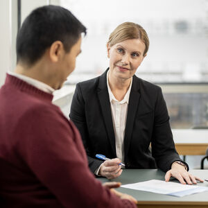 Opiskelija hymyilee ja keskustelee opettajan kanssa pöydän ääressä. Pöydällä on tietokone. Opiskelija katsoo hymyilevä opettajaa, jolla on kynä kädessä. Pöydällä on paperi ja tietokone.