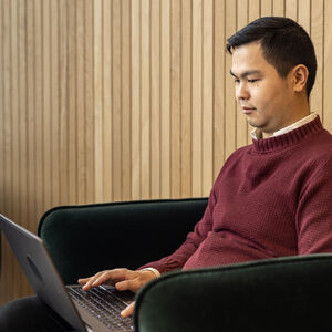 En person i en röd tröja sitter i en stol och arbetar på en bärbar dator. Träpanelevägg i bakgrunden.