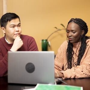 A woman and a man are sitting at a table. There is a notebook and a computer on the table.
