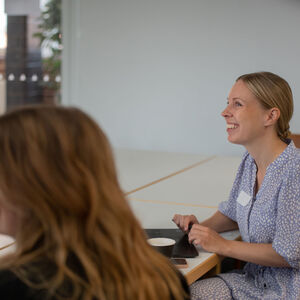 Kaksi ihmistä istuu pöydän ääressä ja katsovat hymyillen eteenpäin. Pöydällä on tietokoneita.