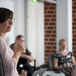 Nainen seisoo ja katsoo sivulle. Taustalla on muita ihmisiä.