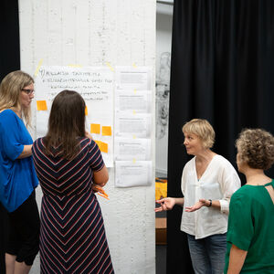 Neljä henkilöä keskustelee seinän vieressä. Seinällä on suuri paperi, jossa on muistiinpanoja ja muistilappuja. å ett stort papper på väggen bredvid finns frågeställningar skrivna med tusch. 