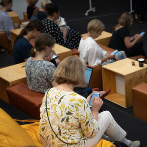 Yleisössä istuu ihmisiä jotka katsovat puhelimiaan.