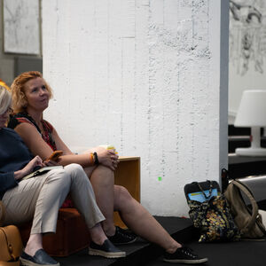 kaksi yleisössä istuvaa naista. Toinen heistä katsoo eteenpäin ja toinen puhelinta.