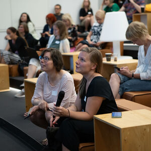 Ihmisiä istuu yleisössä. Eturivissä istuvalla naisella on mikrofoni kädessä.