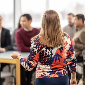 A person in a colourful patterned sweater is presenting to a group of people, some of whom are seated at a table with a laptop.