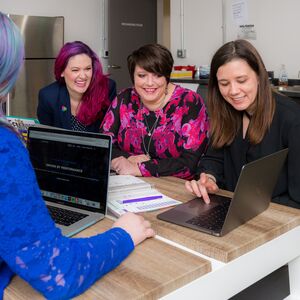 Neljän hengen ryhmä istuu puisella pöydällä kannettavien tietokoneiden kanssa, keskustelee ja tekee muistiinpanoja.