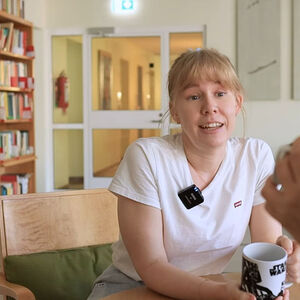 Two people having a conversation at a table, holding mugs. Bookshelves and a hallway are visible in the background.