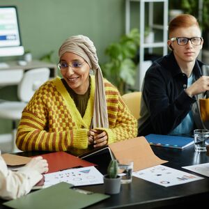 Ryhmä ihmisiä kokouksessa pöydän ympärillä, jossa on kannettavia tietokoneita, asiakirjoja ja vesilaseja vihreässä huoneessa.