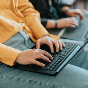 Close-up of two people typing on laptops. One wears a yellow shirt and jeans. Their hands and keyboards are visible.