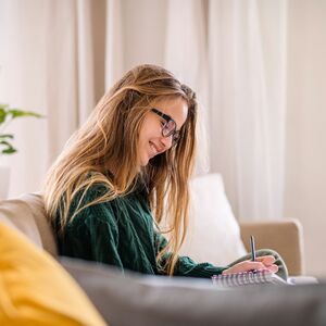 Person med långt hår skriver i en anteckningsbok, sittande på en soffa med en gul kudde, växt och dekorationer i bakgrunden.