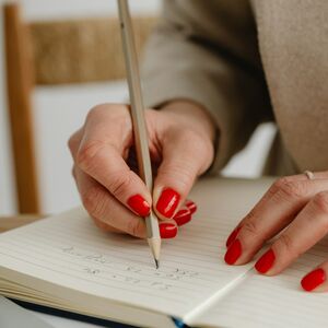 Henkilö, jolla on punaista kynsilakkaa, kirjoittaa muistikirjaan, yllään beige bleiseri.
