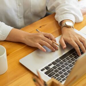 En person skriver på en bärbar dator med en penna i handen, på ett träbord. Det finns en vit mugg i närheten.