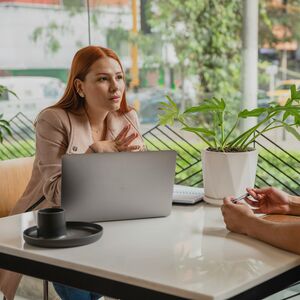 Kaksi ihmistä keskustelee pöydän ääressä, jossa on kannettava tietokone, vihko ja ruukkukasvi toimistossa tai kahvilassa.