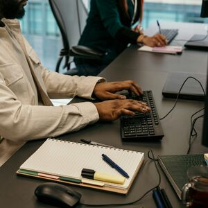 Två personer arbetar vid skrivbord med tangentbord. En har anteckningsbok med pennor, mus och dryck i närheten.
