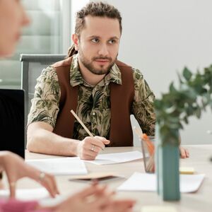 Ryhmä ihmisiä kokouksessa pöydän ääressä papereiden ja kynien kanssa. Yhdellä on vihreä kuviollinen paita ja ruskea liivi.