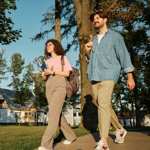 Two people walking on a sunny day, carrying bags and books, surrounded by trees and buildings in the background.