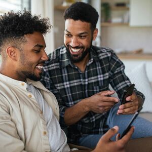 Kaksi ihmistä istuu sohvalla puhelinta katsoen. Toinen on pukeutunut beigeen paitaan, toinen ruudulliseen paitaan.