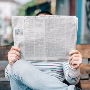 A person in a striped shirt sits on a bench, reading a newspaper that covers their face.