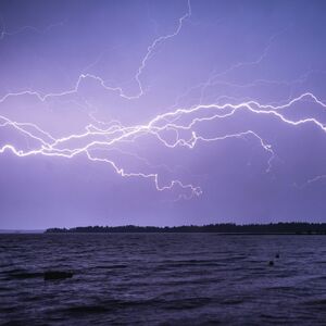 Bright lightning bolts illuminate a dark sky above a large body of water with a distant tree line on the horizon.