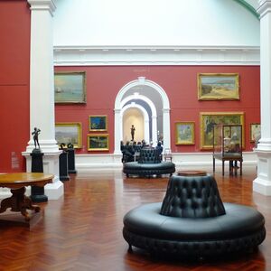 Konstgalleri med röda väggar, målningar, skulpturer och svarta lädersäten i mitten.