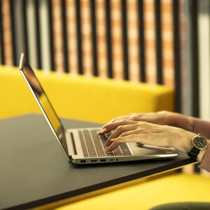 Henkilö vaaleanpunaisessa neuleessa kirjoittaa kannettavalla tietokoneella keltaisilla kalusteilla olevassa työtilassa.