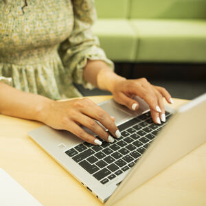 Henkilö kirjoittaa kannettavalla tietokoneella puupöydällä, päällään vihreä kukkamekko pitkine hihoineen.