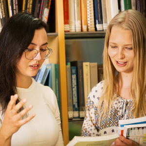 Kaksi henkilöä kirjastossa, toinen elehtii kädellään ja toinen pitää kirjaa. Taustalla on kirjahyllyjä.