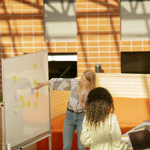 A group of people having a meeting in front of a whiteboard with sticky notes on it. They are in a bright room with large windows.