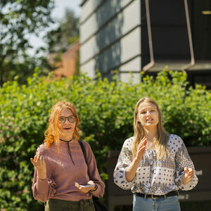 Två personer står utomhus nära grönska, en i rosa tröja och den andra i mönstrad vit tröja.