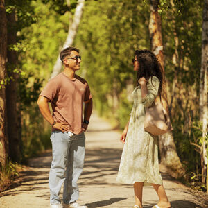 Kaksi ihmistä juttelee aurinkoisella metsäpolulla, toisella on ruskeapaita ja farkut, toisella vaalea mekko ja sandaalit.