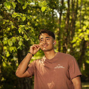A person wearing a brown T-shirt stands in a forested area, with their hand touching a leafy branch.