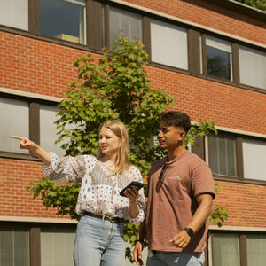 Kaksi henkilöä seisoo lähellä bussipysäkin kylttiä, yksi osoittaa ja toinen pitelee puhelinta. Taustalla on tiilirakennus.