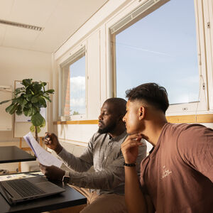 Kaksi ihmistä huoneessa, jossa on suuret ikkunat. Toinen pitelee papereita ja toinen katsoo niitä. Pöydällä on kannettava tietokone.