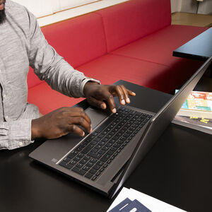 Mies kirjoittaa kannettavalla tietokoneella mustan pöydän ääressä, taustalla punainen sohva.