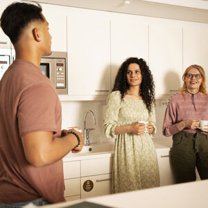 Kolme ihmistä seisoo työpaikan keittiössä pitäen mukeja, taustalla on pesuallas ja mikroaaltouuni.