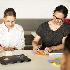 Två personer sitter vid ett bord och skriver på klisterlappar med en bärbar dator och en telefon på bordet.