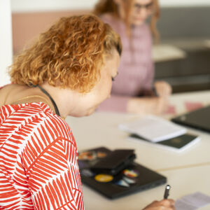Kolme ihmistä pöydän ääressä kannettavien tietokoneiden ja muistikirjojen kanssa. Tarkennettu nainen on kiharatukkainen ja punavalkoisessa paidassa.