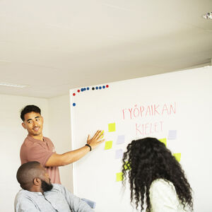 Mies esittelee muistilappuja valkotaululla kolmelle istuvalle ihmiselle. Taululla lukee 'TYÖPAIKAN KIELET'.