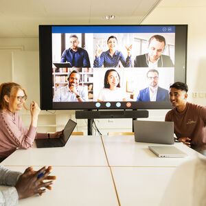 Ihmisiä kokoushuoneessa osallistumassa videoneuvotteluun. Suuri näyttö näyttää lisää ihmisiä puhelussa.