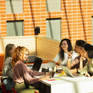 Ryhmä ihmisiä istuu pöydän ääressä, jossa on kannettavia ja muistiinpanoja, tiiliseinän äärellä kirkkaassa sisätilassa.