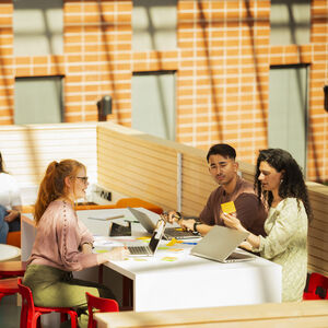 A group of people working at a table with laptops and notes, while others sit on a sofa with laptops in a bright, modern space.