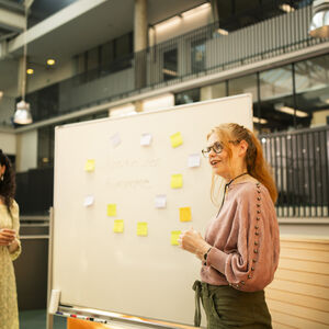 Kaksi henkilöä seisoo valkotaulun edessä, joka on täynnä muistilappuja, modernissa sisätilassa.