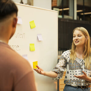 Kaksi henkilöä keskustelemassa ulkona, valkotaululla on muistilappuja. Nainen elehtii käsillään.
