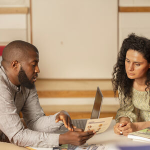 Kaksi ihmistä istuu pöydän ääressä, jossa on kannettava tietokone ja kirjoja. He keskustelavat paperista.